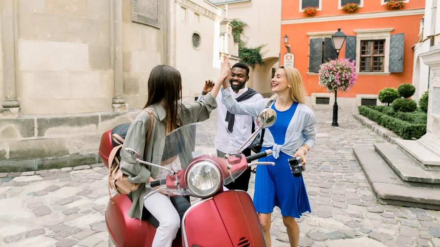 Three happy, multiracial people giving a high five in Rome, with one woman sitting on a Vespa.