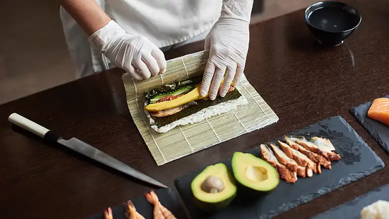 Chef preparing rolling sushi with fresh ingredients on a bamboo mat.