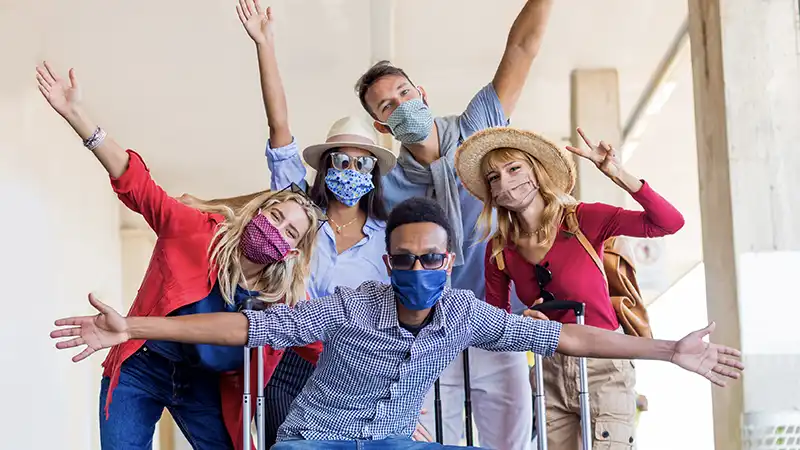 Multiracial friends with masks at a train station, symbolizing safe travel practices and the question of whether travel insurance is necessary.