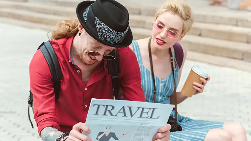 Stylish couple reading a travel newspaper and discussing if travel insurance is necessary while enjoying coffee to go.